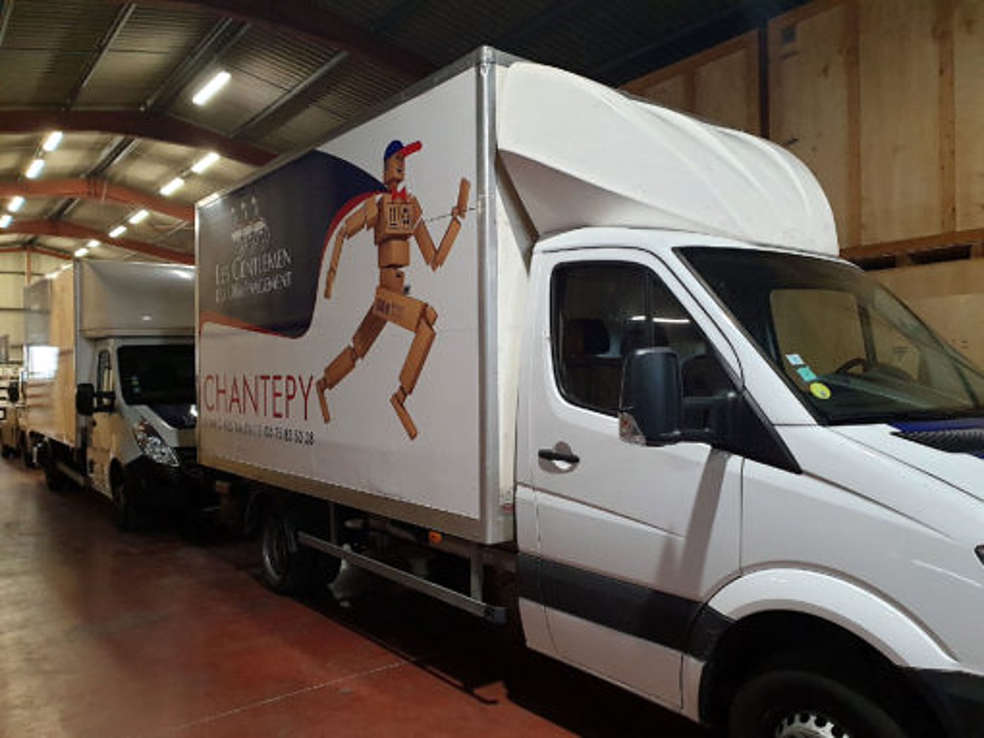 Deux camions de déménagement blancs stationnés dans un entrepôt, celui du premier plan arborant un mannequin en bois et le nom Chantepy.