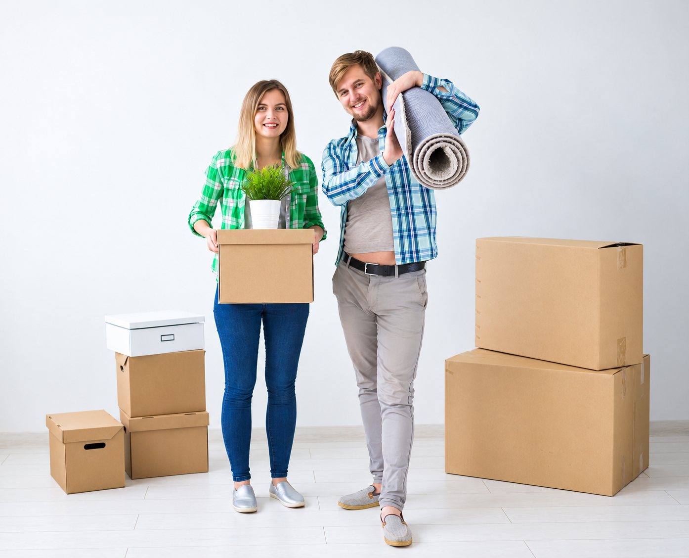Un jeune couple souriant tient une plante dans un carton et un tapis roulé, entouré de cartons de déménagement dans une pièce blanche.