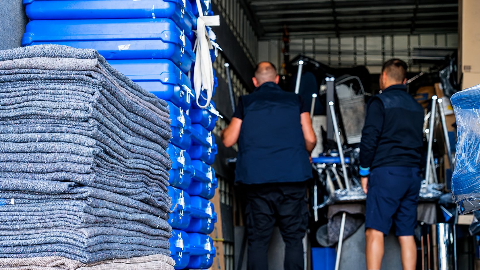 Deux déménageurs vus de dos chargent du mobilier dans un camion, à côté d’une pile de couvertures de protection grises et de caisses bleues empilées.