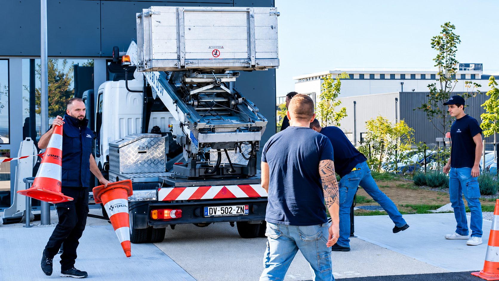 Des hommes placent des cônes de signalisation autour d’un camion équipé d’un monte-meubles sur un parking devant un bâtiment moderne.