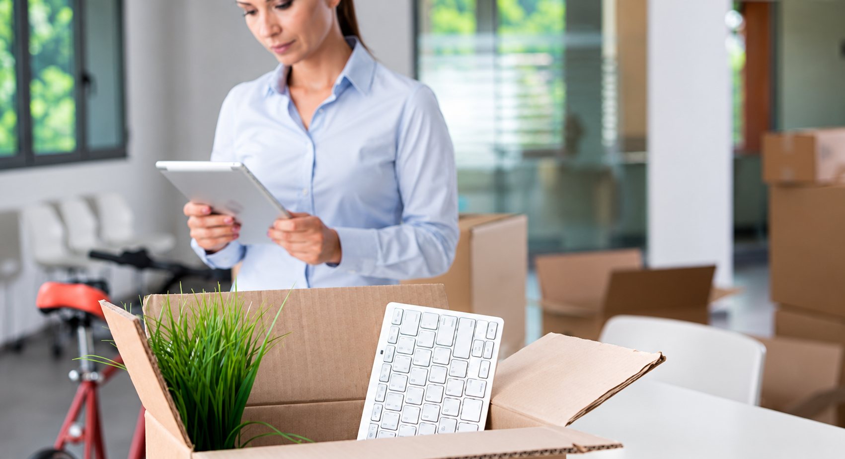 Une femme en chemise bleue tient une tablette dans un bureau, avec au premier plan un carton contenant un clavier et une plante verte.