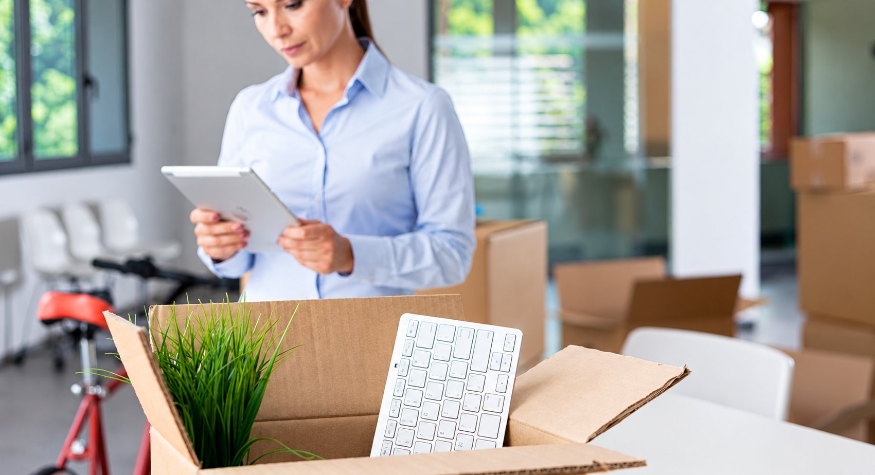 Une femme en chemise bleue regarde une tablette derrière un carton ouvert contenant un clavier blanc et une plante, dans un bureau en plein déménagement.