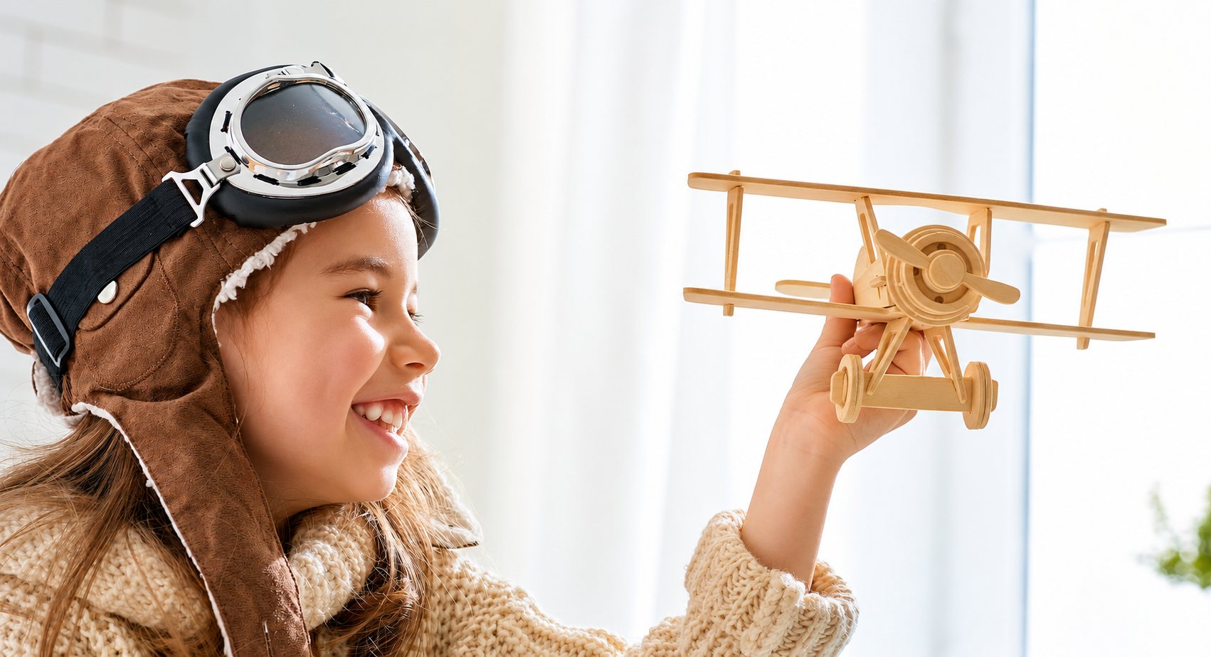 Une petite fille souriante portant un casque et des lunettes d’aviateur tient un avion biplan en bois clair devant une fenêtre lumineuse.